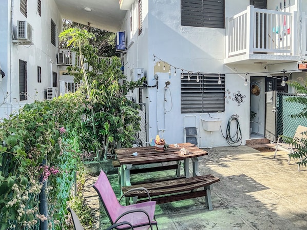 A courtyard with a wooden table and benches, surrounded by greenery and a white building with multiple windows and air conditioning units.