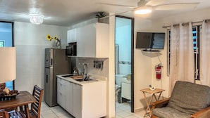 Una cocina compacta con microondas, lavamanos y armarios, un comedor con mesa y sillas, y un televisor en la pared.