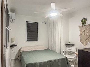 A bedroom with a bed, a ceiling fan, a window with blinds, a wall-mounted air conditioner, a wall decoration, and a desk with a chair.