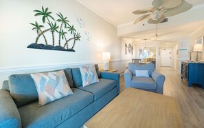 A living room with a sofa, armchair, and coffee table. There is a ceiling fan, a wall-mounted shelf with palm trees, and a dining area in the background. - Phoenix I 1143 2 Bedroom Condo (Orange Beach)