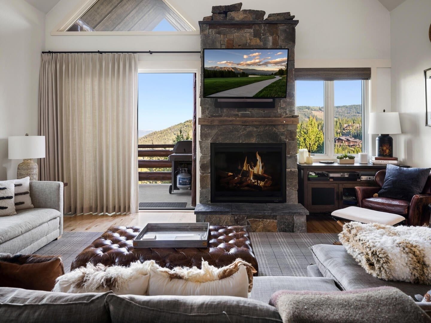 A living room with a stone fireplace, a flat-screen TV mounted above it, and a view of a mountainous landscape through large windows.