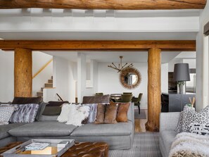 A modern living room with a grey sectional sofa, wooden beams, a dining area with a round mirror, and a staircase. - Saddle Ridge S2 3 Bedroom Home (Big Sky)