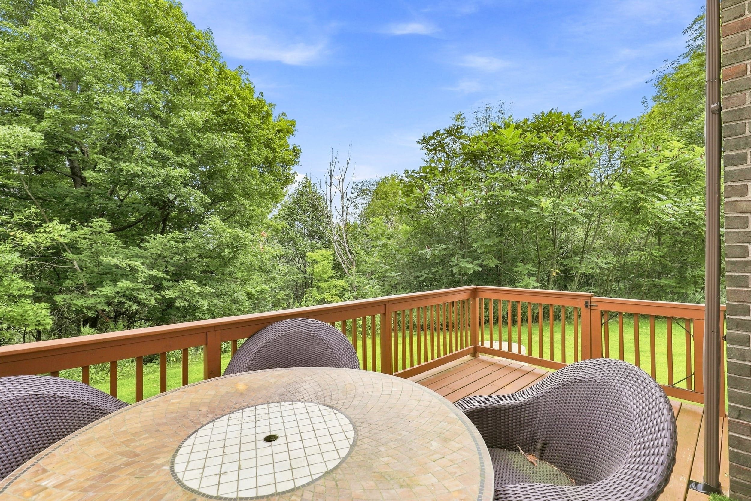 A deck with a round table and wicker chairs, surrounded by a wooden railing, overlooking a lush green landscape.