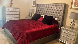 Een slaapkamer met een geborduurde hoofdbord, een rode spreideken, twee nachtkastjes met lampen, een dressoir en een ingelijste foto aan de muur.