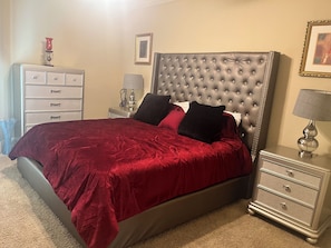 Un dormitorio con cabecera capitonada, colcha roja, dos mesitas de noche con lámparas, cómoda y un cuadro enmarcado en la pared.