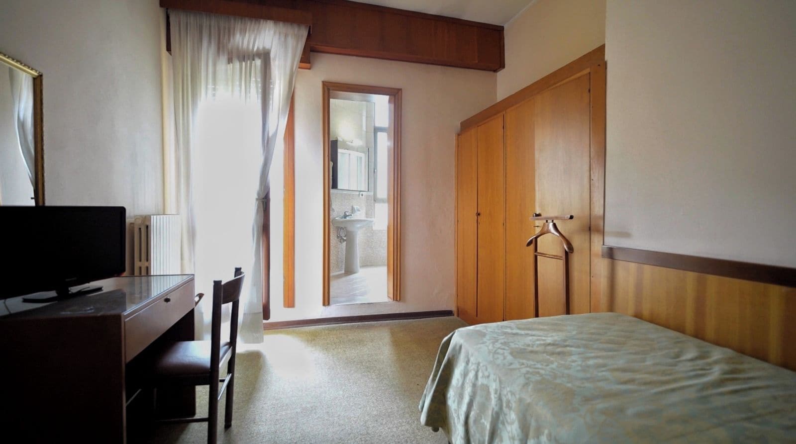 Ein Hotelzimmer mit einem Bett, einem Schreibtisch mit Fernseher, einem Sessel, einem Kleiderschrank und einem durchgehenden Türspalt erkennbaren Badezimmer.