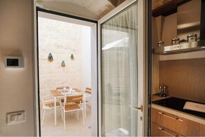 A modern kitchen with a black countertop and wooden cabinets, a dining area with a table and chairs, and a view of a brick wall through a glass door. - A Stay in the Village: Corte Suite – Village Charm with a Private Courtyard (Ceglie Messapica)