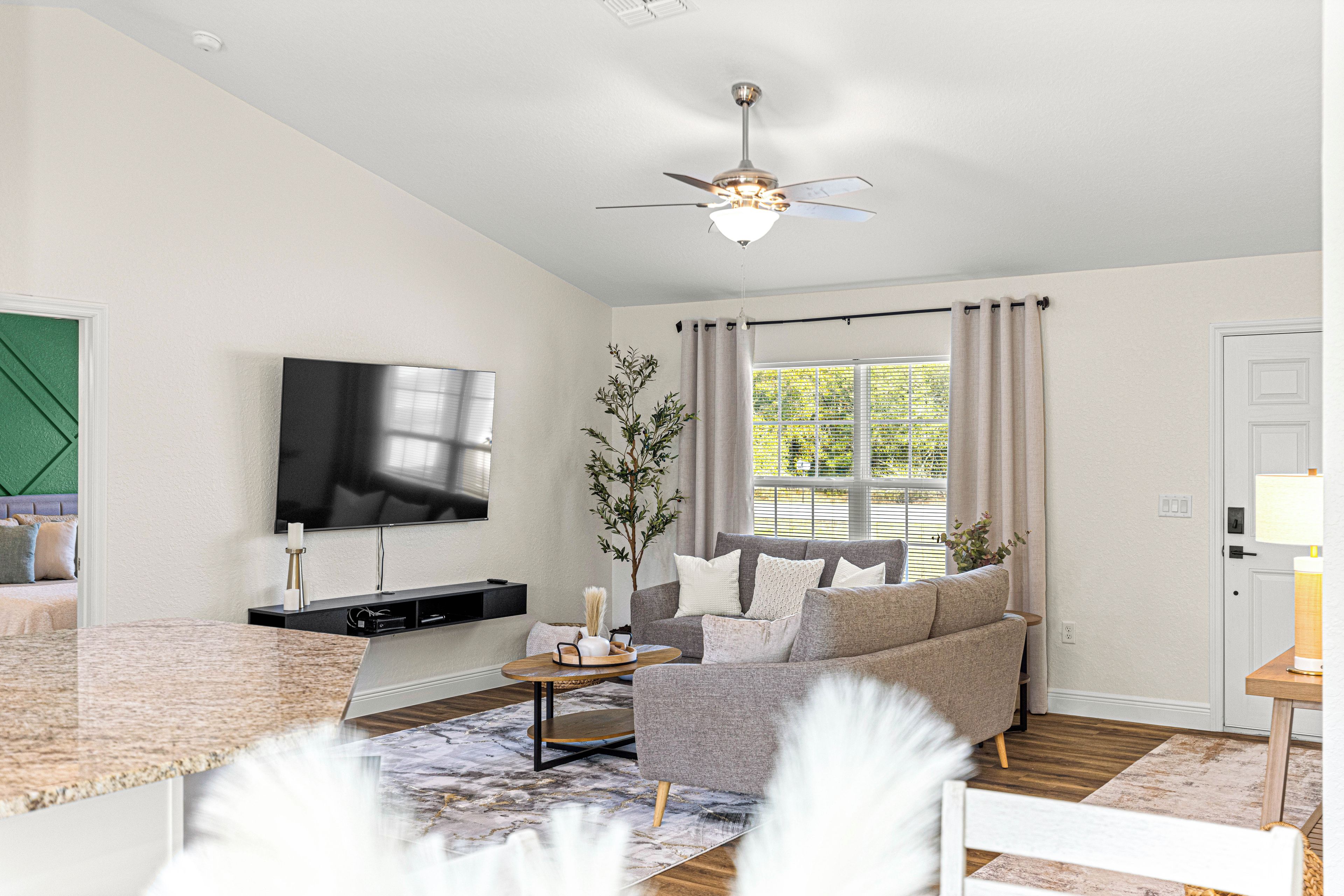 A modern living room with a flat-screen TV, a grey sofa, a wooden coffee table, and a ceiling fan.