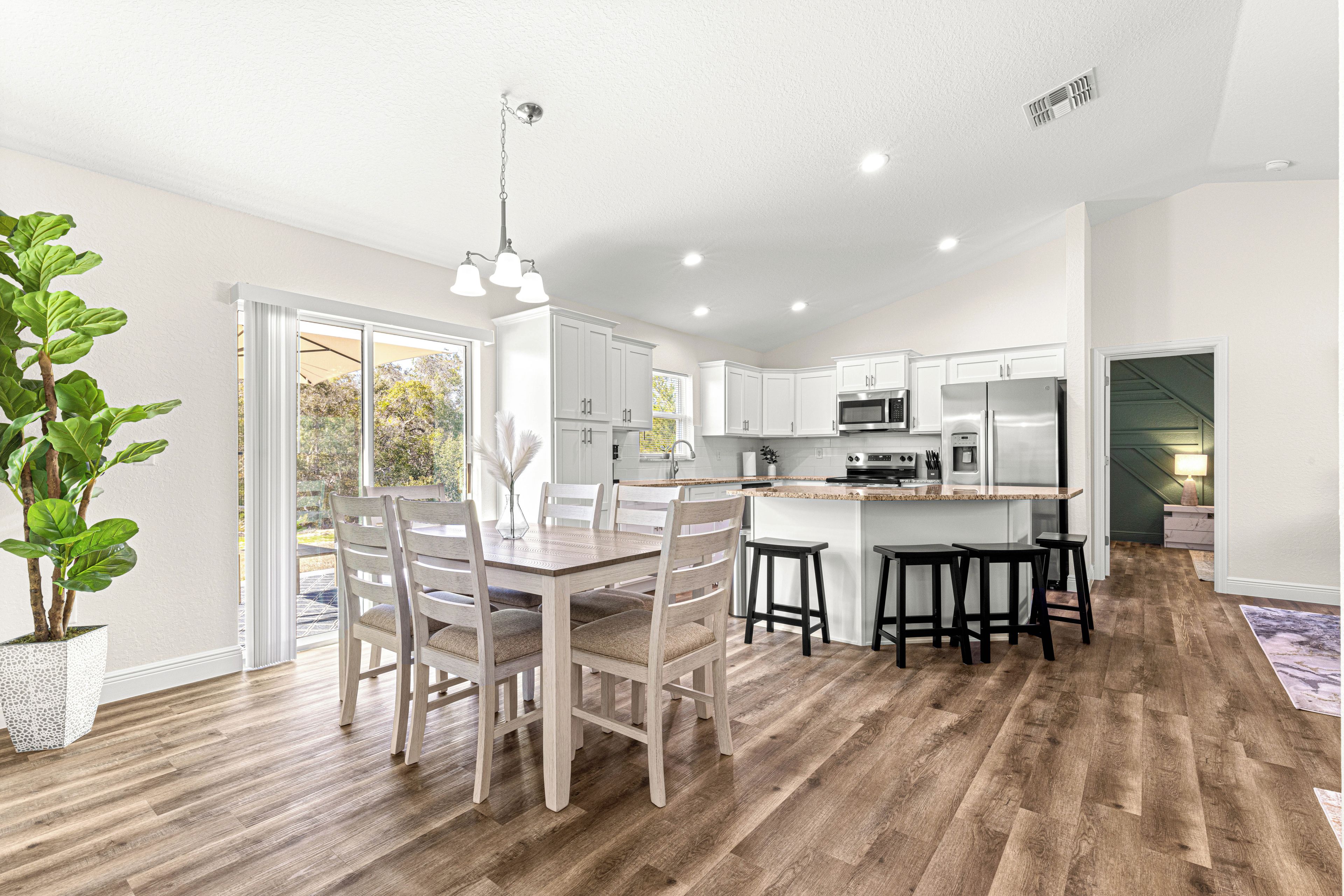 A modern kitchen with white cabinets, a central island with bar stools, and a dining area with a table and chairs.