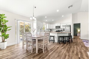 A modern kitchen with white cabinets, a central island with bar stools, and a dining area with a table and chairs. - Spacious 4-bedroom home, minutes from I-75 with a game room and sleeping for 8! (Ocala)