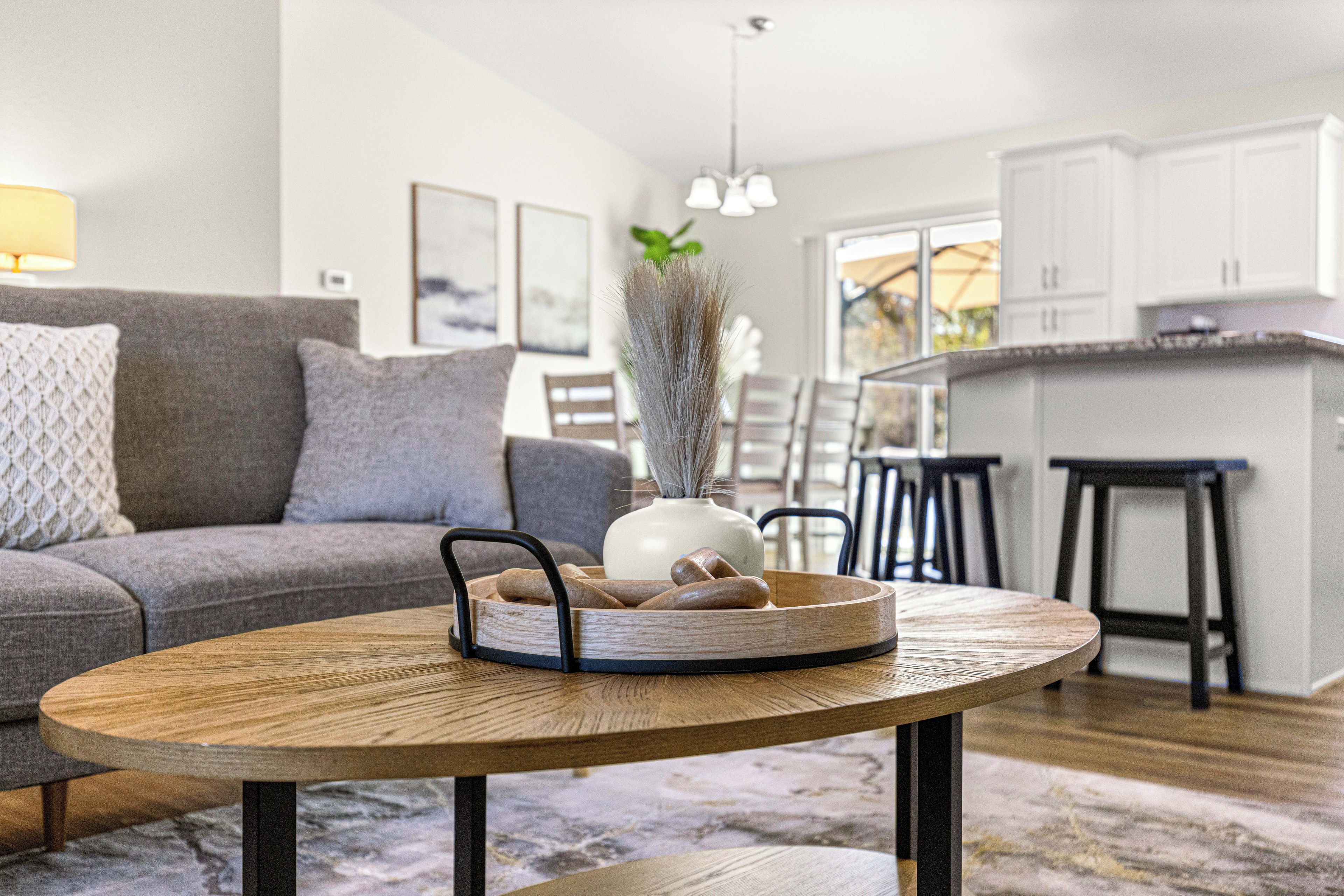 A modern living room with a grey sofa, a round wooden coffee table with a woven tray, and a vase with dried plants.