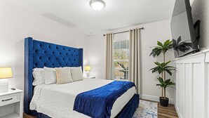 Un dormitorio con una cama grande, una mesita de noche, una lámpara, una ventana con cortinas y una planta en maceta.
