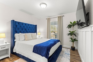 Un dormitorio con una cama grande, una mesita de noche, una lámpara, una ventana con cortinas y una planta en maceta.