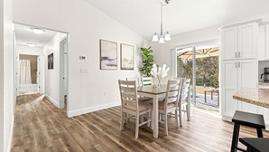 Una cocina amplia con armarios blancos, un comedor con mesa y sillas de madera, y vista al exterior a través de grandes puertas correderas de vidrio.
