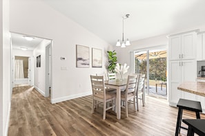 Una cocina amplia con armarios blancos, un comedor con mesa y sillas de madera, y vista al exterior a través de grandes puertas correderas de vidrio.