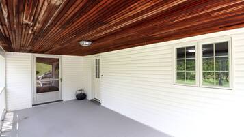 A covered porch with a wooden ceiling, white siding walls, and a door leading outside.