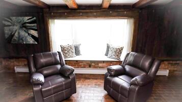 Une pièce avec deux fauteuils en cuir, une grande fenêtre avec des stores, un mur en briques et une poutre en bois au plafond.