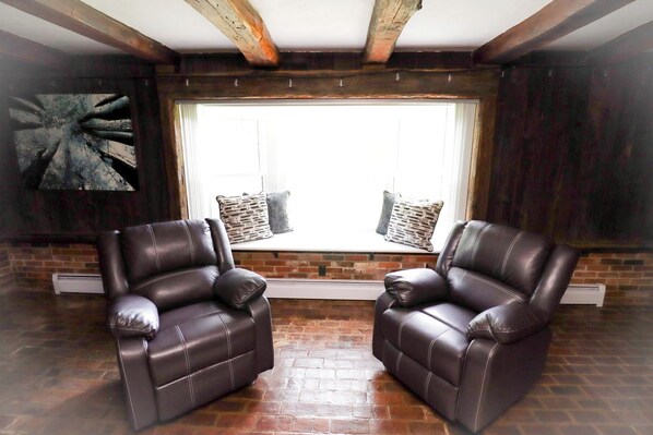 A room with two leather armchairs, a large window with blinds, a brick wall, and a wooden ceiling beam.