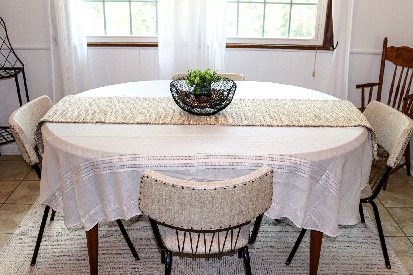 A round dining table with a white tablecloth, a woven placemat, and a small plant centerpiece. There are four chairs with woven seats and backs. The room has a window with white curtains and a wooden chair in the corner.