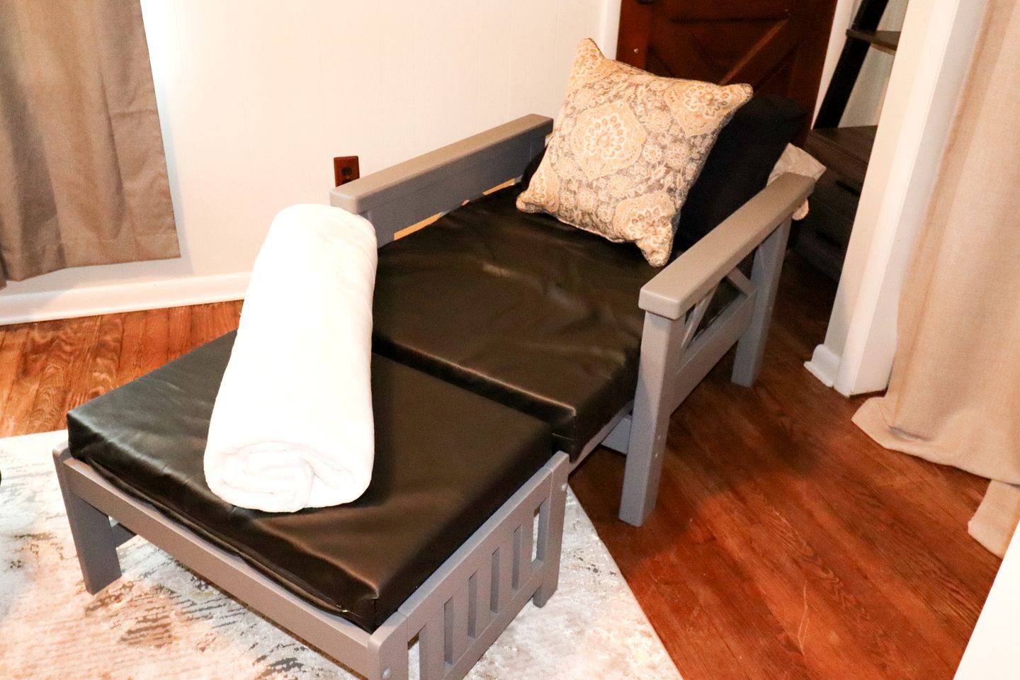 Une petite chambre avec un lit simple, un cadre gris, un matelas noir, une serviette blanche et un oreiller à motifs.