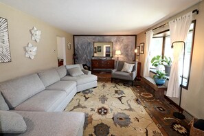 A living room with a sectional sofa, a small armchair, a patterned rug, a mirror, and a window with curtains.