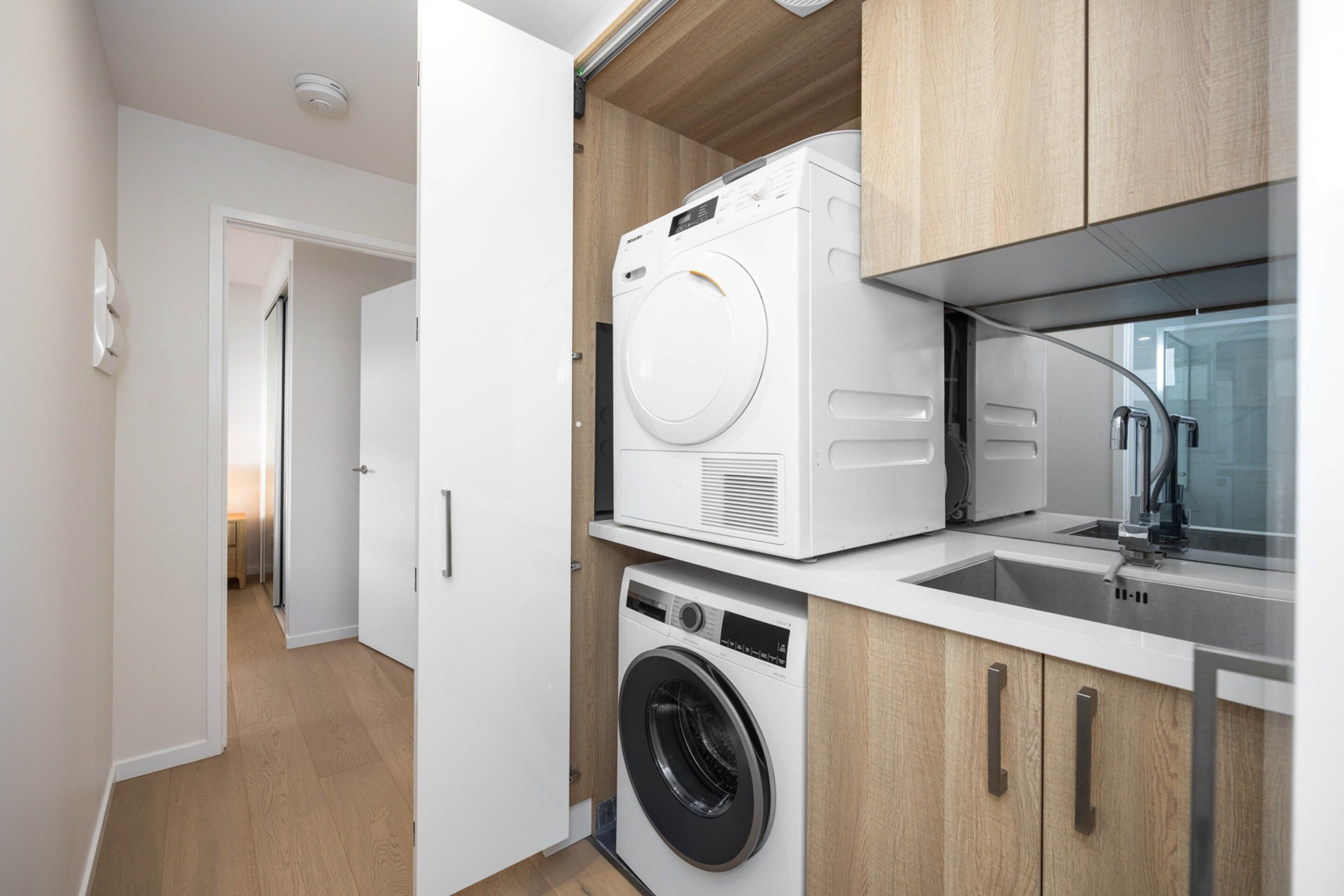 A compact laundry area with a front-loading washer and dryer, wooden cabinetry, and a small sink.