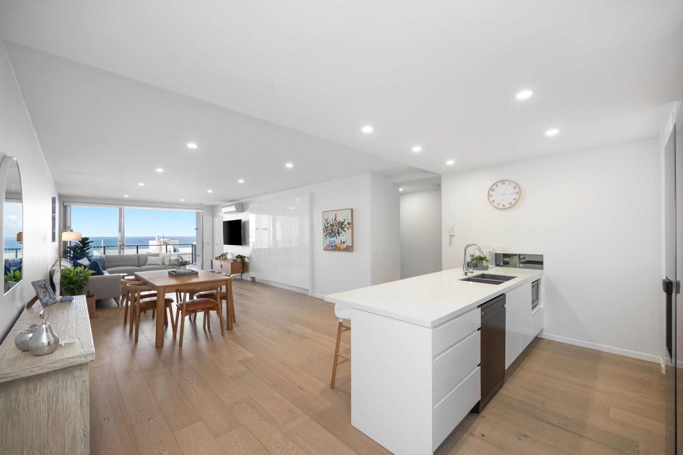 A modern kitchen with a central island, a dining area with a sofa and chairs, and a view of the sea through large windows.