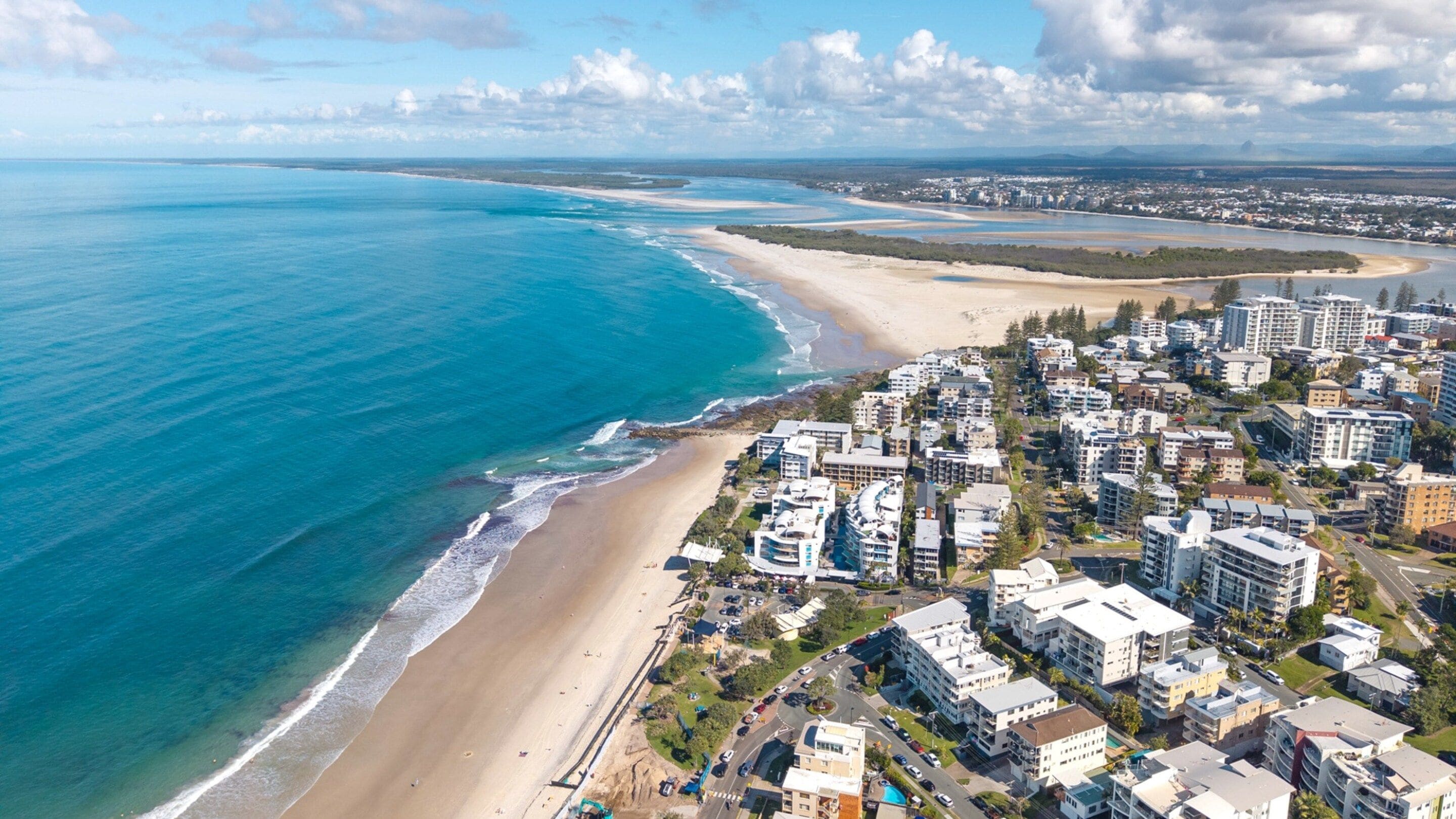 An aerial view of a coastal residential area with beachfront properties, roads, and a distant cityscape.