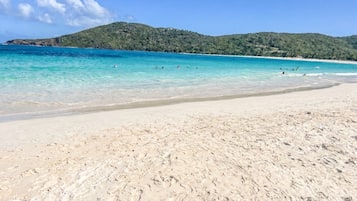 A beach with clear turquoise water, sandy shore, and a hilly landscape in the background.