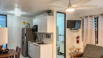 A compact kitchen with a refrigerator, microwave, and sink. There's a dining table with chairs and a small side table. A ceiling fan is visible, and a fire extinguisher is mounted on the wall.