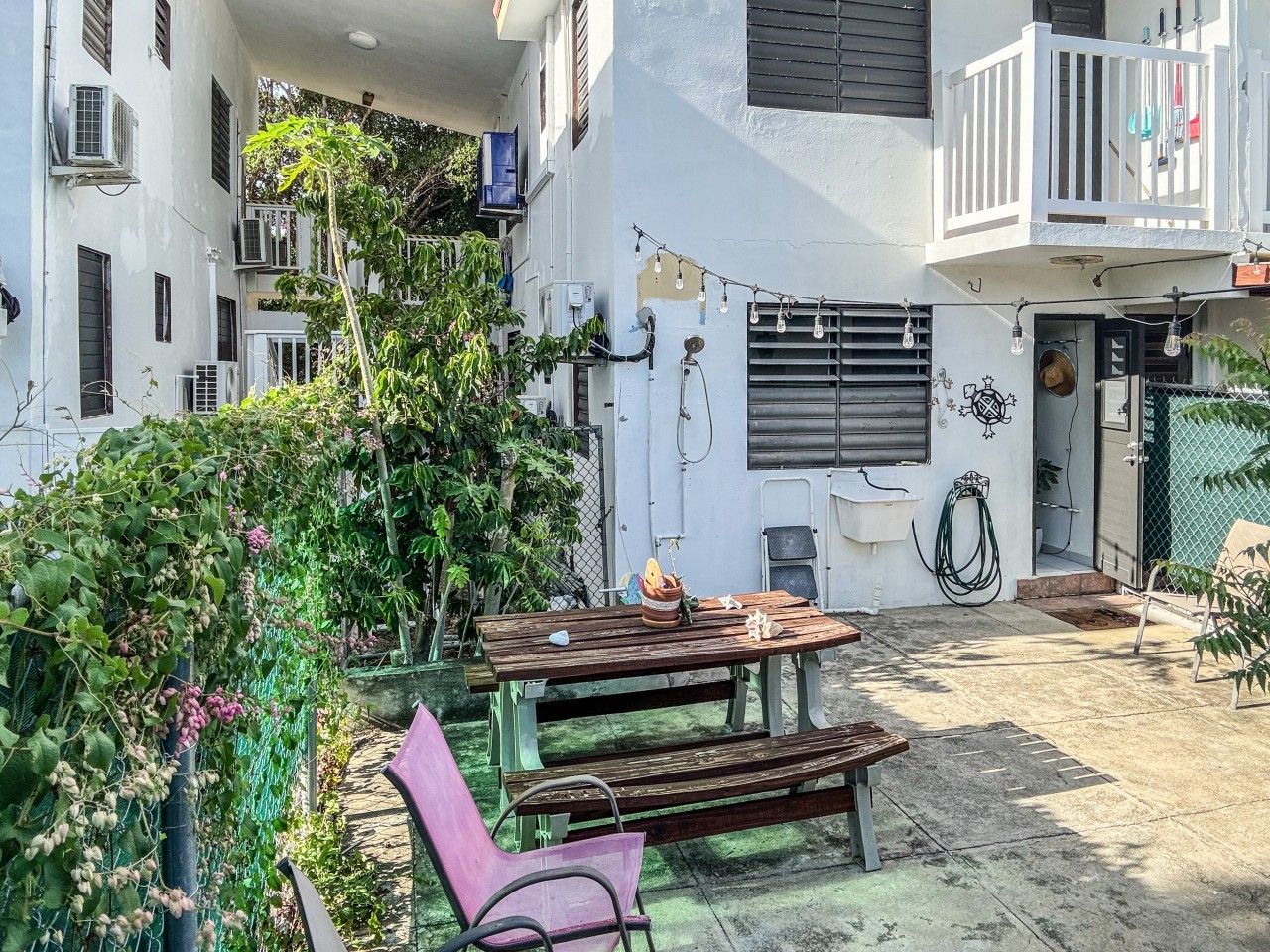 A courtyard with a wooden table and benches, surrounded by greenery and buildings.