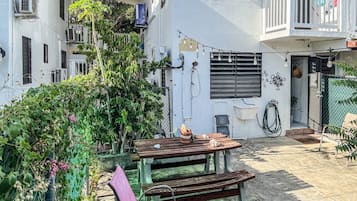 A courtyard with a wooden table and benches, surrounded by greenery and buildings.