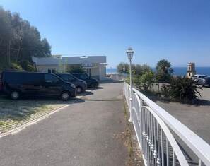 En parkeringsplats med flera bilar, en byggnad med en vit fasad och en tydlig utsikt över havet i bakgrunden.