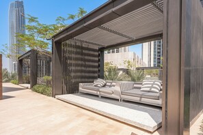 Un área de estar moderna al aire libre con un sofá y cojines, una pérgola y vistas de edificios altos.