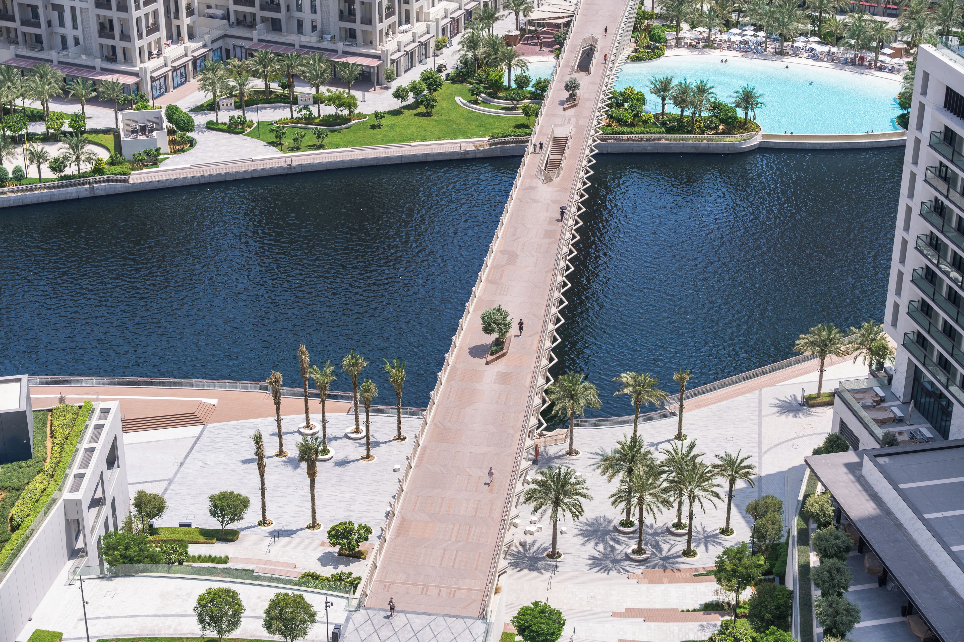 A waterfront promenade with a bridge, palm trees, and a pool area.