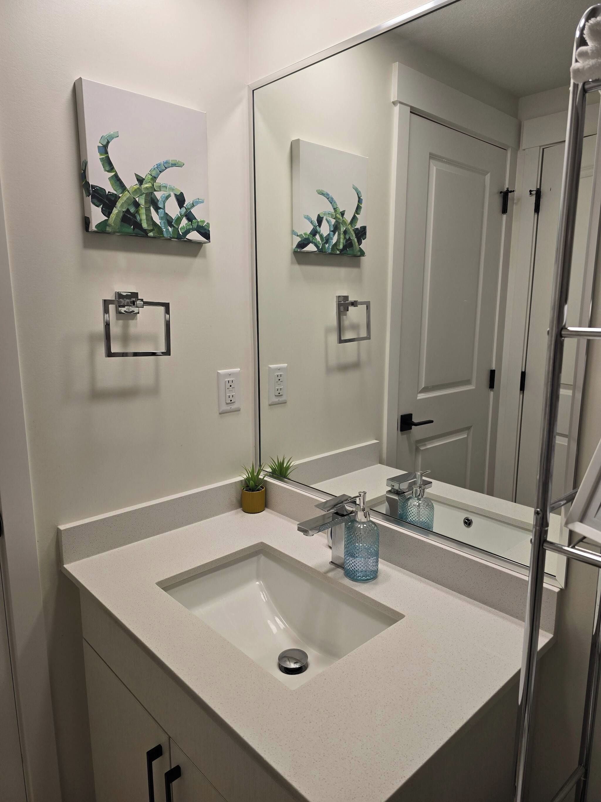 Un baño con un espejo grande, un lavabo, un perchero para toallas y una planta en maceta.