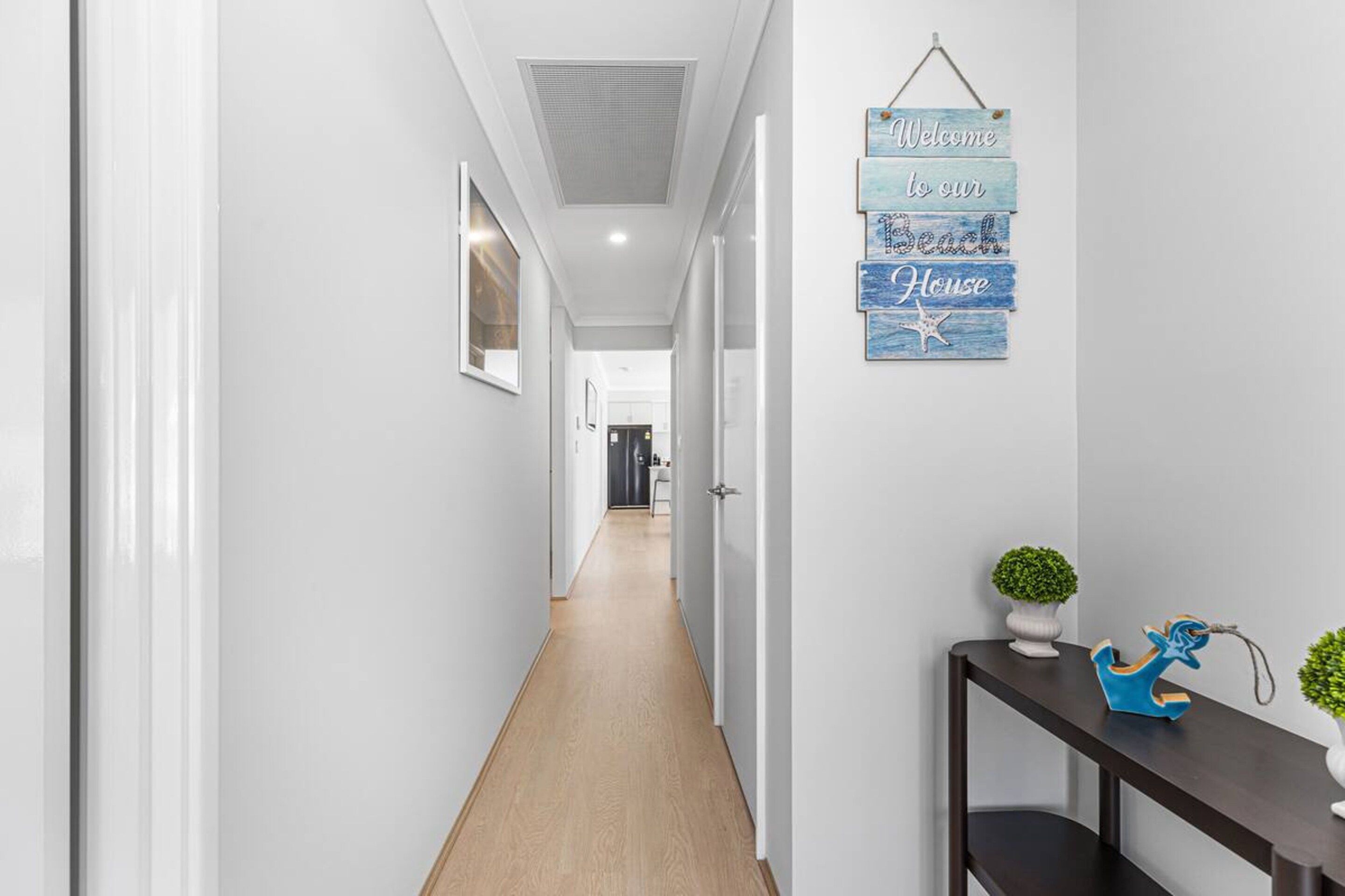 A hallway with a wooden floor, white walls, and a hanging sign that reads 'Welcome to our Beach House.'