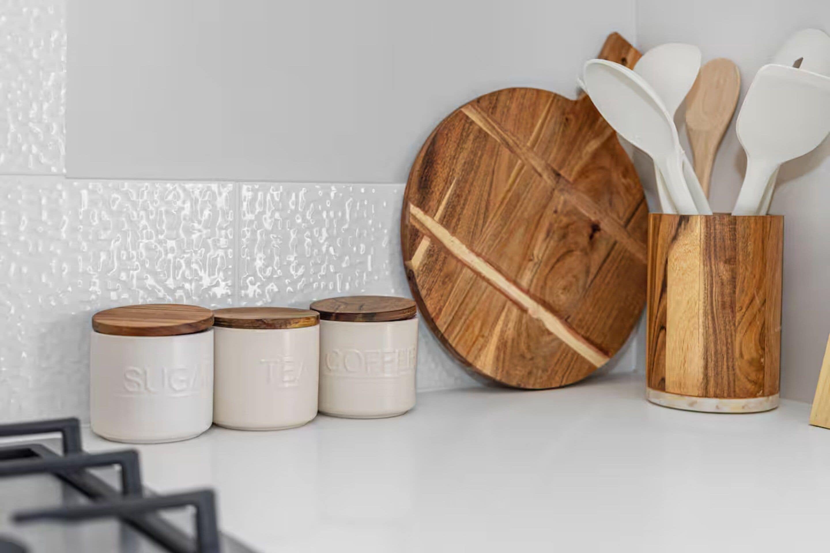 Three white ceramic containers with wooden lids, a wooden cutting board, and a wooden utensil holder with white utensils.