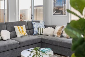 A grey sectional sofa with multiple pillows, a white coffee table with books and a plant, and a view of the outdoors through large windows. - Alkimos Coastal Escape (Alkimos)