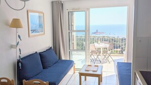 Een kamer met een bank, een eettafel, stoelen, een kitchenette en een balkon met uitzicht op zee.