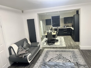 Una sala de estar moderna con un sofá, un comedor con mesa y sillas, y una cocina con armarios oscuros.