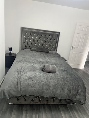 A bed with a tufted headboard, a rolled towel, and a bedside table with a lamp.