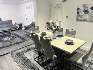 A dining area with a table set for four, surrounded by grey upholstered chairs. A vase with flowers is placed on the table, and there are two doors in the background.