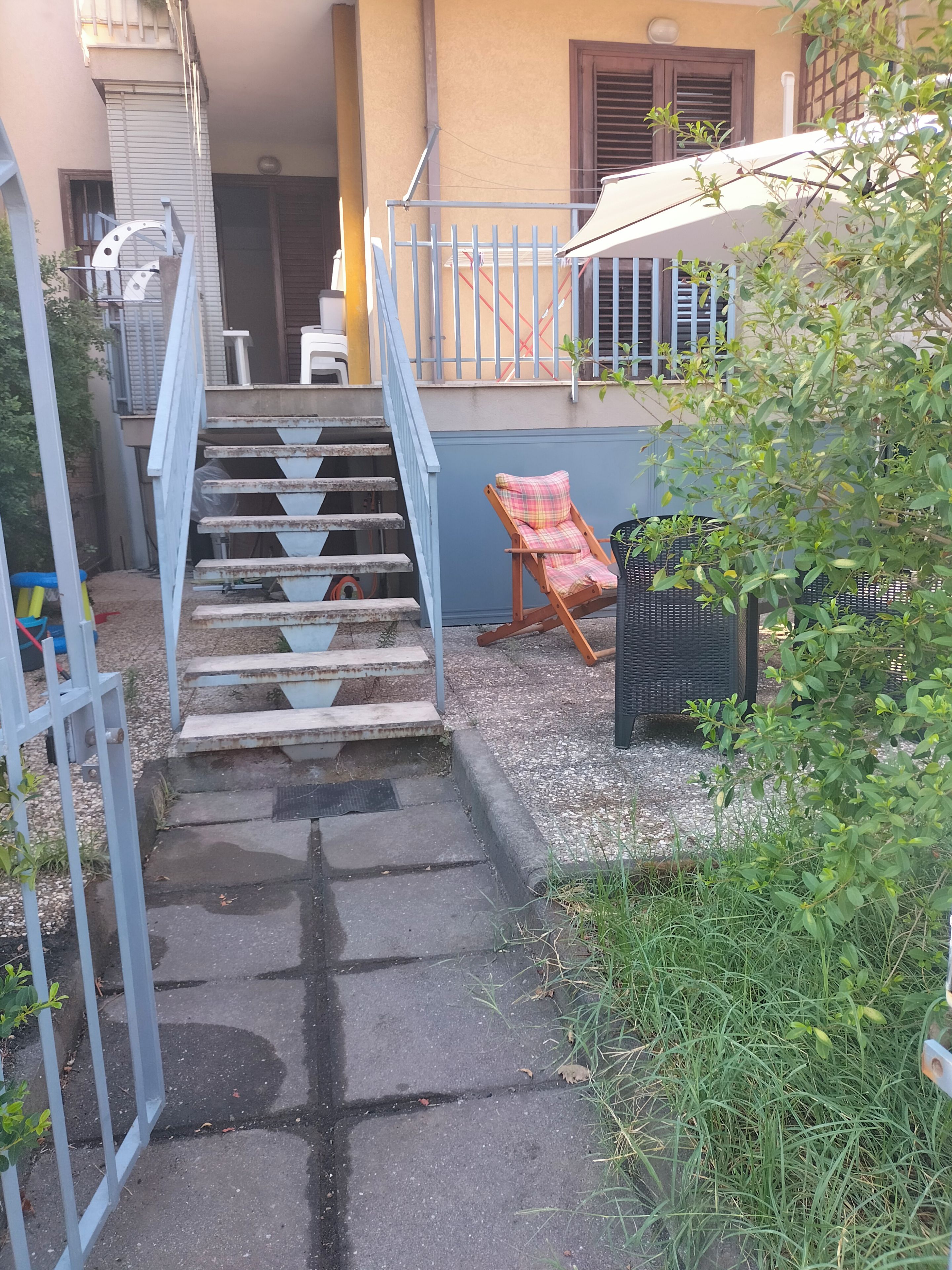 Un escalier menant à un bâtiment doté d’une rambarde bleue, d’une chaise de patio et d’un petit espace jardin.