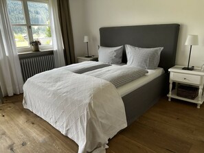 Ein ordentlich bezogenes Bett mit einer gesteppten weißen Daunendecke, karierten Kissen und einem grauen Kopfteil. Zwei Nachttische mit Lampen und ein kleiner Korb sind vorhanden. Durch ein Fenster mit Vorhängen ist ein Blick auf Bäume und ein Haus zu sehen.
