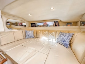 Schiffsinnenraum mit einer beige gepolsterten Sitzgelegenheit, einem Fenster mit Vorhängen und einem Regal mit verschiedenen Gegenständen.