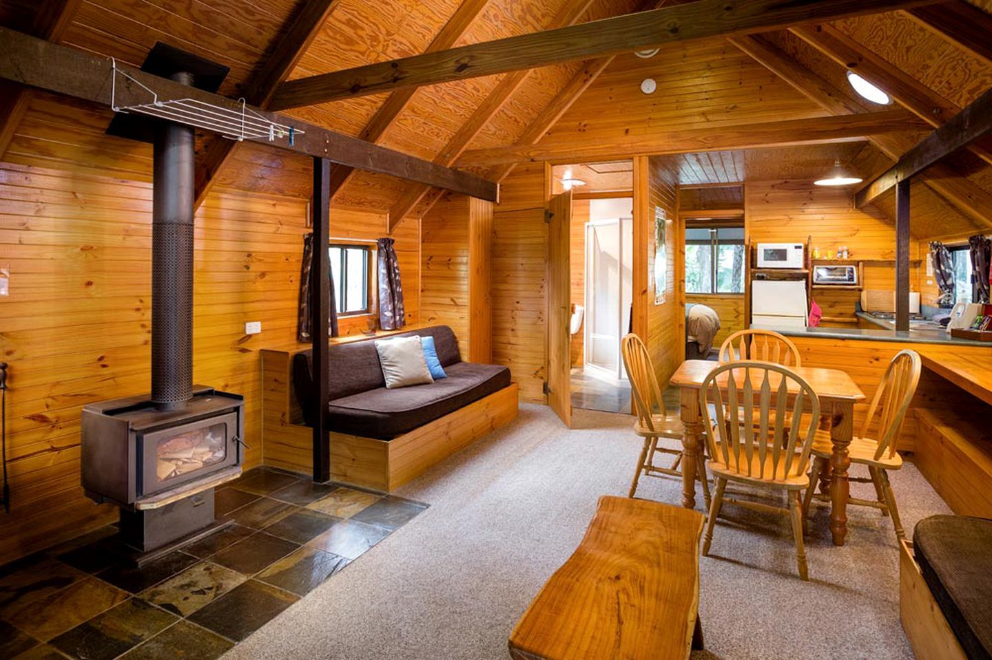 A cozy cabin interior with a wood-burning stove, a sofa, a dining table with chairs, and a kitchen area.