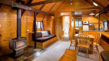 A cozy cabin interior with a wood-burning stove, a sofa, a dining table with chairs, and a kitchen area.