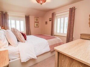 A bedroom with a bed, a bedside table, a window with curtains, and a wall-mounted light fixture. - Myrtle Cottage (Lynmouth)