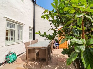 A patio with a glass-top table and wicker chairs, a hose reel, and a white building with windows. - Myrtle Cottage (Lynmouth)